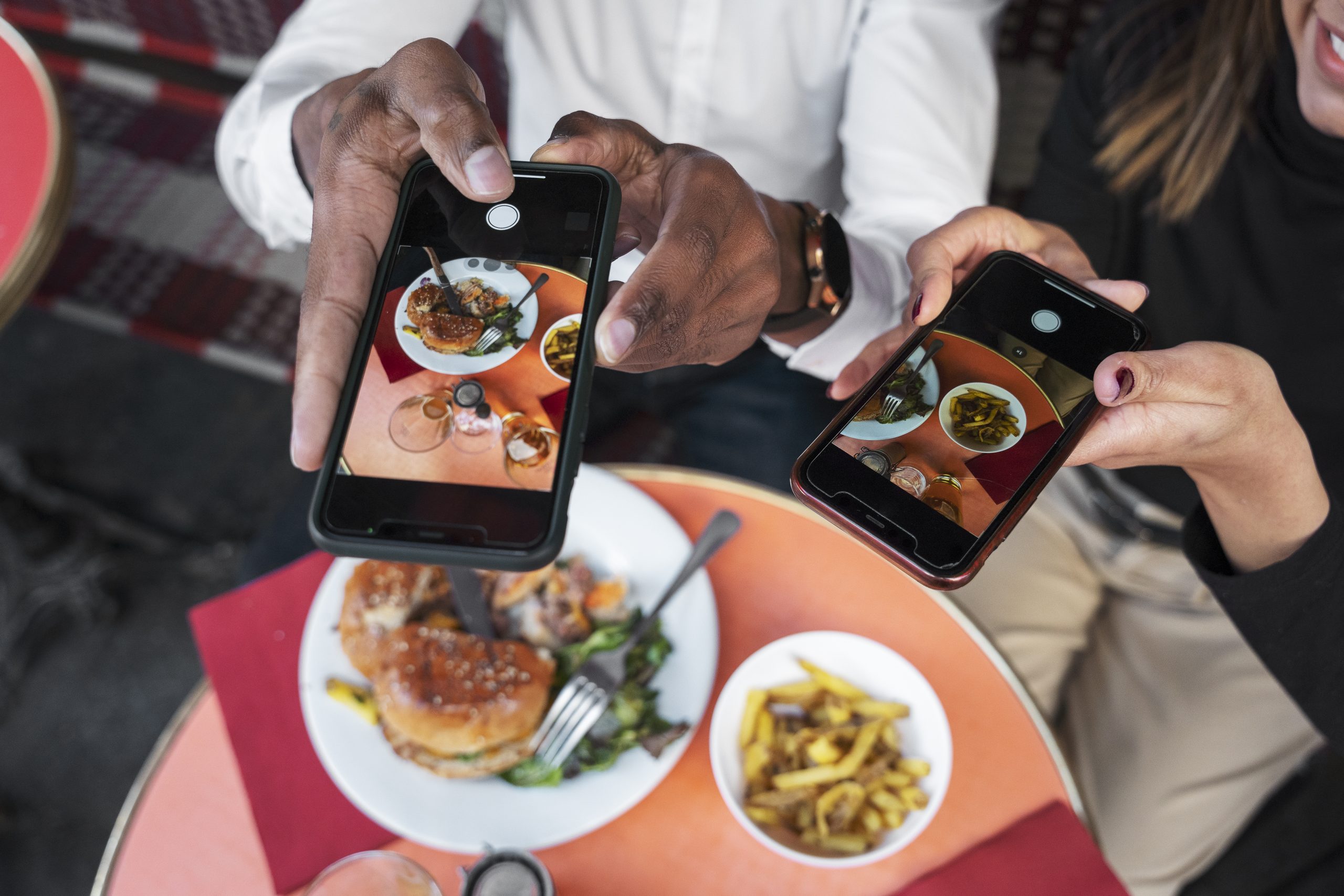 Two people photographing their meals—burgers, fries, salads—with smartphones at a restaurant, capturing content for social media. Highlights the everyday moment of creating shareable food content while building an Instagram account from scratch.
